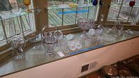 Wide-angle of glass servingware collection on window sill, showing diverse items including bowls, pitchers, decanters, and glasses.