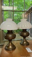 Photo of both hurricane lamps together on a wooden surface by a window, shows differences in glass shade styles and lamps' bases.