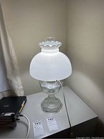 Clear glass lamp with white frosted hurricane shade on a white surface.