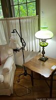 Photo showing both the floor lamp and the vintage hurricane-style table lamp side by side in a room environment, the floor lamp has a black metal stand with a white ruffled fabric shade, the table lamp is on a brown wooden table with a green glass base and frosted white glass shade.
