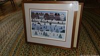 Full view of one framed art print resting on rug, showing winter ice skating scene with houses in background