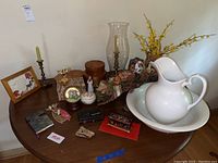 Wide view of table showing ironstone pitcher and basin, two candlesticks with green candles, faux yellow flowers, snow globe, covered box, decorative frame, and several small decorative items.