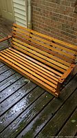 Wooden porch swing showing full seat and armrests with chains attached, placed on wooden deck near brick wall.