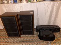 Photo showing two Royal 4A bookshelf speakers with wooden finish and black fabric grills alongside two Sony stereo boom boxes, one gray and one black.