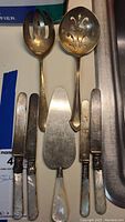Overview of sterling cutters with mother of pearl style handles and two large sterling serving utensils showing tarnish and need for cleaning.