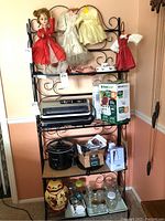 Front view of the black metal bakers rack loaded with various items including dolls on the top shelf, kitchen appliances, and jars on lower shelves.