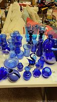 Full view of assorted cobalt blue glass items arranged on table, showing swan, pitchers, vases, glasses, small bottles, and bowl.