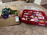 Two containers of assorted vintage Christmas ornaments on carpet: one red plastic organizer with multiple ornament types, and one cardboard box with additional decorations.