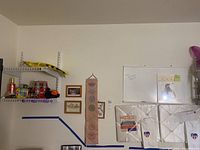 Photo showing corner of room with shelf holding Coca Cola collectible cans, other small items, and a section of wall with whiteboard and framed memorabilia.