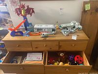 Wide shot of dresser top with Hess vehicles including RV, tractor trailer, race car, motorcycle, and helicopter; Hot Wheels raceway track parts with red dragon topper; open drawers show plush toys and Nerf balls.