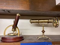 Two vintage kaleidoscopes on shelf, one wooden with brass and one all brass with stand.