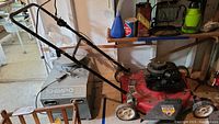 Photo showing the full red Briggs & Stratton lawn mower with handlebar and wheels in a garage setting, revealing dirt and some rust.