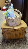 Smaller woven basket on top of Longaberger picnic basket containing sealed plastic bag with yellow plastic cutlery and other picnic items, shown on carpet floor.