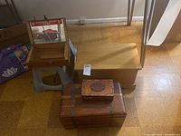 Overview photo showing three wooden boxes stacked: small cedar chest on top, medium wood storage box beneath, and mirrored display box partly open to show inside text. Also shows plastic step stool in background.