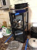 Photo of black shelving unit with stacked Revere Ware pots and pans including roasting pans and baking pans. Cooling racks visible on floor.