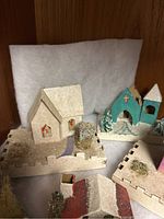 Single close-up photo of three Putz houses: white house on castle-edge base, blue church with snowy base, and another castle-edge base with moss/tree.