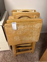 Nine wooden tray tables stacked folded upright showing worn tops and folding legs.