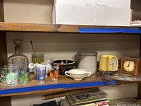 Shelf displaying vintage clocks, drinking glasses, ceramic crock, decorative teacup, and glass decanter.