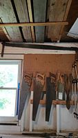 Four vintage hand saws with wooden handles hanging on a wooden rack below a large wooden plank ceiling, showing aged wood and metal blades