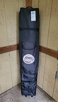 Quik Shade black rolling carrying case vertically placed against a wall with a front zippered pocket and a visible logo patch.