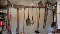 Wide view of various yard tools hanging on a textured wall including brooms, rakes, shovels, and a shop light