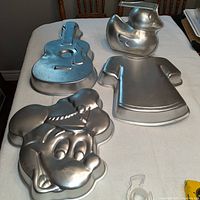 Photo showing four aluminum cake pans shaped like a guitar, Mickey Mouse, a duck, and a shirt on a table.
