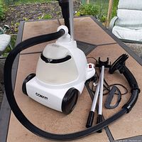White Conair fabric steamer with clear water tank and black flexible hose. Multiple accessories including two metal rods, brush, and fabric clip are placed beside it on a table outdoors.