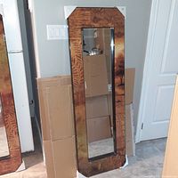 Floor mirror with a wood grain finish frame and protective corner covers leaning against a wall near a door and folded cardboard boxes.