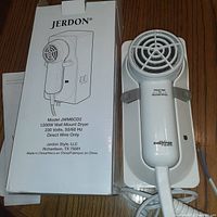 Box and wall mount hair dryer placed on a wooden surface. Dryer is white with a circular vented grill and a handle attached to a white plastic mounting plate. Blue and brown wires extend from the base for hardwiring.