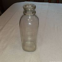 Photo shows a single clear glass milk bottle on a light-colored tablecloth, capturing the shape and clarity of the bottle.