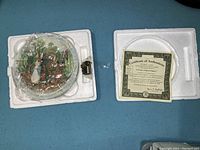 Collector plates in protective styrofoam packaging alongside certificates authenticating the plates, showing one plate wrapped with visible characters and floral motifs