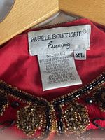 Label of red beaded jacket showing Papelle Boutique Evening brand and fabric details.