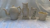 Group photo of four Irish Belleek porcelain items showing teapot, owl vase, harp figurine with shamrocks, and flower pot on a light background