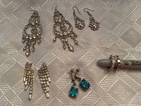 Five pairs of rhinestone earrings arranged on a textured metallic surface, showing a variety of shapes, sizes, and colors for pierced ears.