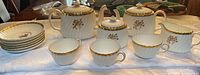 Full tea set with all pieces displayed on a white cloth, showing the 2 teapots, sugar bowl with lid, creamer, 3 cups, and 6 saucers decorated with gold rose design.