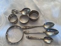 Photo of entire lot showing spoons, napkin rings, and bracelet arranged on cloth background.