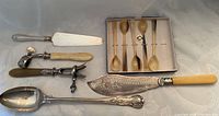Full view of serving pieces laid out on table including stuffing spoon, cake server, fish slice, lamb leg holders, and egg spoons in box.