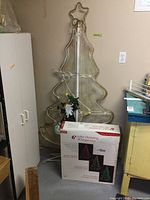 Metal framed 6 ft rope light Christmas tree without lights on, leaning against wall with original box visible in front.