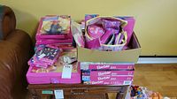 Tabletop display showing Barbie puzzles, dolls, accessories in a box, and clothing items stacked on an end table alongside leather furniture.