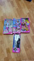 Photo showing all four dolls in original packaging, three Barbie dolls and one Ken doll, on wooden floor.