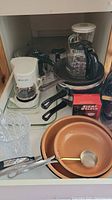 Kitchen drawer containing white coffee maker with glass pot, a glass vase, three brown pans with a metal strainer spoon inside, and a red Eight O'Clock box.
