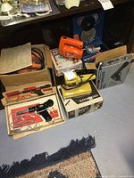 Lot of assorted boxed and unboxed power tools arranged on floor including a soldering iron, sander, cordless drill, and jigsaws.