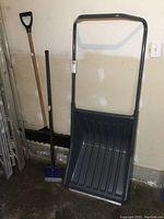 Three snow and ice removal tools including a large black snow scoop with metal handle, a wooden handle snow shovel with black plastic D-grip, and a blue/black ice chipper tool. Tools are standing upright against a beige wall beside folded metal ladders.