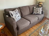 Side and angled view of three-seat brown fabric sofa with two patterned throw cushions on it, showing armrests and seat cushions.