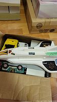 Photo showing the Fisher Price space shuttle inside a cardboard box with visible parts of toy cars beneath it and some Lego components.