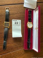 Two wristwatches on a wooden surface, one in a red presentation box with 'Never forget 1945 to 2005' inscription, both with leather bands.