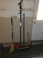 Full view of all yard tools standing against a wall: push broom, electric hedge trimmer, whisk broom, axe, and telescopic pole pruner.