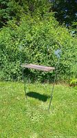 Image showing both green metal shepherd's hooks with attached wooden platforms, standing on grass in front of shrubbery.