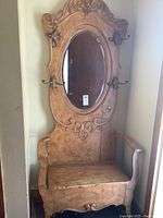 Front full view of painted wooden hall tree with oval mirror, four metal hooks, seat with storage and armrests, showing overall condition and wear.