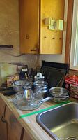 Overview of the kitchen counter with metal baking trays, mixing bowls, cooling racks, and various kitchen utensils stacked and arranged near a sink.